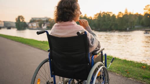 Finanzielle Gefahren bei langer Krankheit - und wie sie sich eindämmen lassen Eine ältere Frau sitzt im Rollstuhl und blickt auf den Fluss.