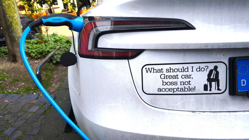 Ein Elektroautomobil der Marke Tesla wird an einer Ladestation aufgeladen. Der Besitzer hat einen Aufkleber an seinem Fahrzeug angebracht. Der Inhalt des Textes besagt das er findet ein grossartiges Auto zu besitzen, aber den Chef der Marke Tesla Elon Musk für nicht akzeptabel hält
