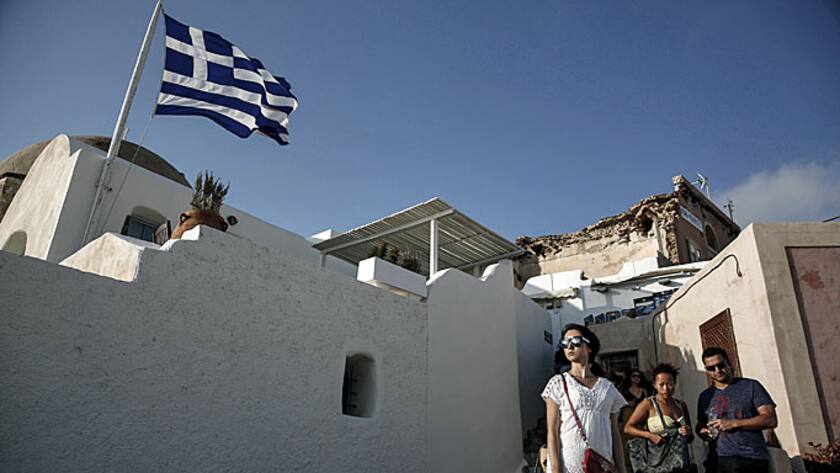 Tourists stand and look at the view beside a Greek national flag flying above a house in Oia village on the island of Santorini, Greece, on Wednesday, May 20, 2015.